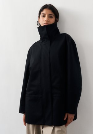 Vrouw draagt een oversized zwarte jas met hoge kraag, staande tegen een effen witte muur, beige broek zichtbaar onder de zoom van de jas.