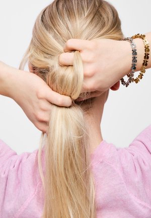 Persona rubia con camiseta rosa recogiendo el cabello largo en una coleta baja, mostrando las manos y pulseras plateadas y doradas con forma de mariposa.