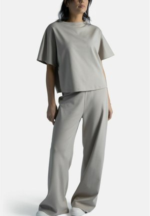 Nainen, joka pukeutuneena löysään beigeen lyhythihaiseen paitaan ja leveälahkeisiin housuihin, seisoo käsi rentona sivullaan yksiväristä taustaa vasten.