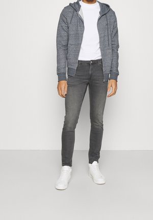 Sweat à capuche gris à fermeture éclair avec cordons, porté sur un T-shirt blanc, associé à un jean slim gris foncé et des baskets blanches. Tissu lisse et coupe décontractée.