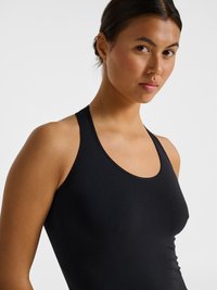 Young woman with dark hair tied back, wearing a fitted black sleeveless athletic top, looking slightly toward the camera against a plain background.