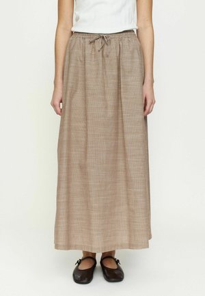 Kvinne iført en beige lang skjørt med snøring i midjen, hvit ermeløs topp og mørkebrune flate sko, stående mot en ensfarget bakgrunn.