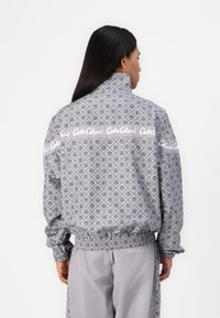 Grijze windbreaker met een diamantpatroon en vredessymbolen, hoge kraag, elastische manchetten en een gebrandmerkte witte tailleband. Bijpassende grijze broek.