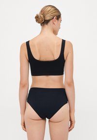 Woman with hair in a bun wearing black lace-trimmed bralette and high-waisted black underwear, facing away against white background.