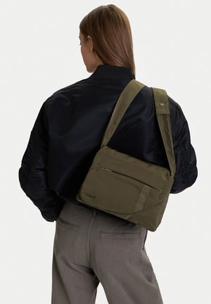 Frau mit schwarzer Jacke und grauer Hose trägt eine olivgrüne Umhängetasche über der Schulter, blickt von der Kamera weg.