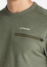 Olivgrön sweatshirt med rund halsringning, med en zip-försedd ficka på vänster bröst och "G-Star Raw"-logotyp i vitt. Slät textur.