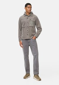 Grijze corduroy jas met een capuchon, voorzien van een borstzak en knoopsluiting. Gecombineerd met grijze broek en beige sneakers.