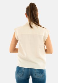 Blusa sin mangas color crema con cuello, fabricada en un tejido suave, líneas limpias y un detalle de yugo en la parte trasera. Combinada con unos jeans azules.