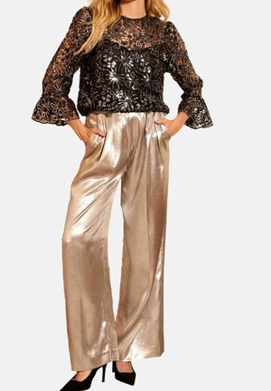 Femme portant un haut en dentelle florale noire avec des manches transparentes et un pantalon large doré brillant, les mains dans les poches, et des talons hauts noirs.