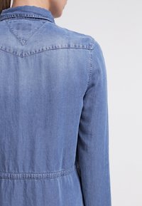 Světle modrá džínsová bunda s špičatým límcem, stáhnutým pasem a zúženým zadním dílem ve tvaru V. Měkká textura s jemnými detaily šití.