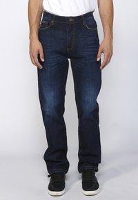 Donkerblauwe denim jeans met een rechte snit, voorzien van gouden stiksels, vijf zakken en een knoopsluiting. Gecombineerd met zwarte sneakers.