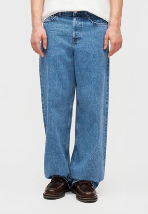 Άτομο που φοράει ανοιχτό μπλε τζιν παντελόνι με φαρδιά πόδια και σκούρα καφέ παπούτσια με κορδόνια, στέκεται μπροστά σε απλό φόντο.