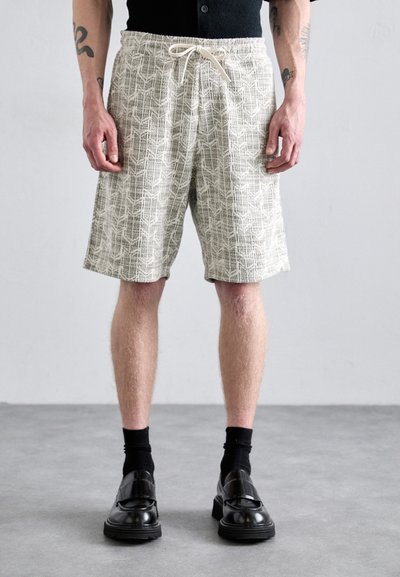 Shorts légers à motifs beiges avec une taille élastique et un cordon de serrage. Portés avec des chaussettes noires et des chaussures en cuir verni.