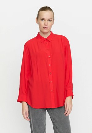 Red long-sleeve button-up shirt with a collar, featuring a loose fit and smooth texture. Paired with gray trousers.