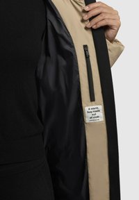 Giacca esterna nera con fodera beige, dotata di una tasca con zip e un'etichetta che recita "Un abbraccio caldo fatto di stoffa." Texture liscia.