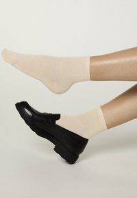 Zapatos planos de cuero negro con acabado liso, combinados con calcetines beige acanalados. El zapato tiene una puntera redondeada y una suela baja y texturizada.