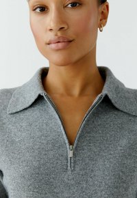 Grijze gebreide pullover met een ritsneklijn en een kraagontwerp. Zachte textuur met een iets getailleerde pasvorm. Rits heeft een metalen trekstang.