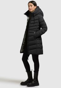 Cappotto piumino nero con cuciture verticali, alto colletto e cappuccio regolabile. Presenta tasche con zip e foderato interno lucido.