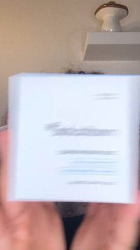 Vit förpackningslåda med blå text och svarta detaljer, hålls i händer med suddig bakgrund. Designen är enkel och rektangulär.