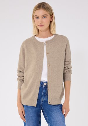 Frau trägt einen beigen, geknöpften Cardigan über einem weißen T-Shirt mit blauen Jeans, steht vor einem schlichten hellen Hintergrund.