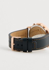 Μαύρη δερμάτινη λουρί με πορτοκαλί ραφές και ροζ χρυσό κούμπωμα, συνδεδεμένη με ροζ χρυσό ρολόι στο χέρι, στραμμένη προς τα κάτω.