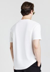 T-shirt bianca in cotone con maniche corte, texture a coste e orlo rotondo, vista dal retro. Design minimalista, senza loghi visibili.