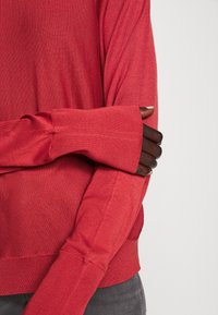 Červený pletený svetr s dlouhými rukávy a žebrovanými manžetami. Tkanina má měkkou strukturu a je zobrazena na šedém denimu.