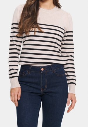 Vrouw met een beige trui met lange mouwen en zwarte horizontale strepen, en donkerblauwe high-waisted jeans, staand met ontspannen armen.