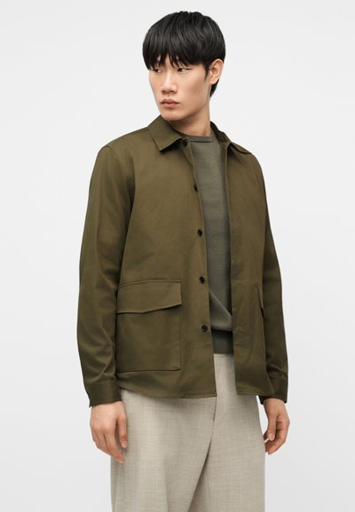 Νεαρός άνδρας με μαύρα μαλλιά, ντυμένος με ελιάς πράσινο κουμπωτό μπουφάν και πουλόβερ, μπεζ παντελόνι, κοιτάζει προς τα αριστερά πάνω σε λευκό φόντο.