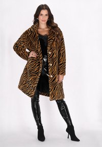Faux bontjas met een tijgerstrepenpatroon in bruin en zwart, met enkele knopen en grote knopen, gecombineerd met glanzende zwarte leggings en enkelboots.