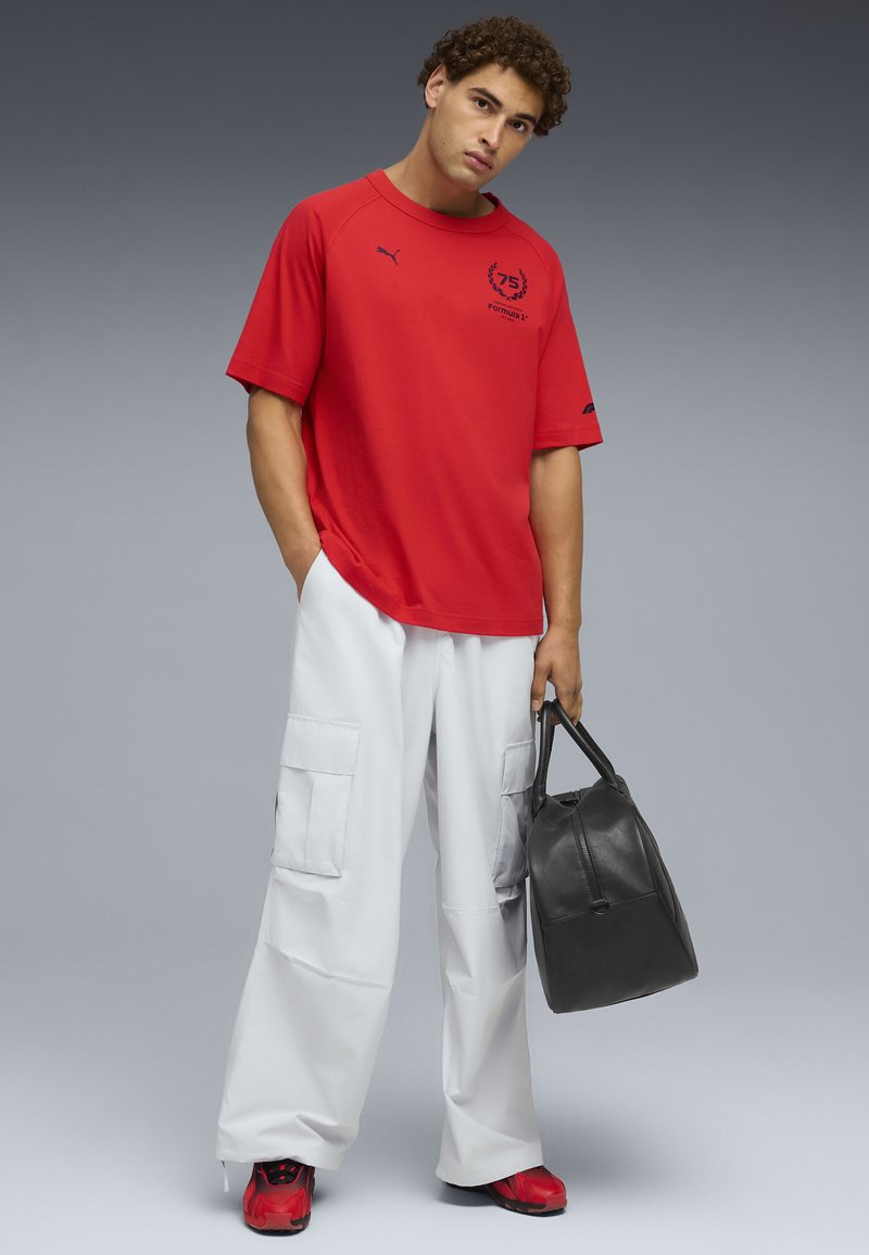 T-shirt rouge à manches courtes avec un logo sur la poitrine, associé à un pantalon cargo blanc avec de grandes poches. Tient un sac en cuir noir.