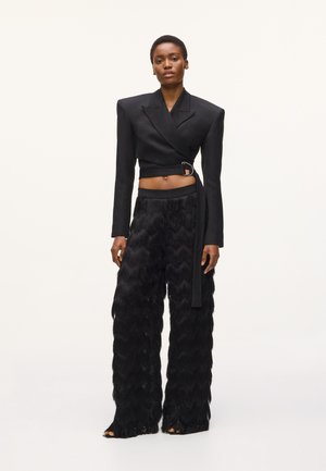 Femme portant un blazer noir court à larges épaules et ceinture à boucle, associé à un pantalon noir large texturé et des chaussures à bouts ouverts.