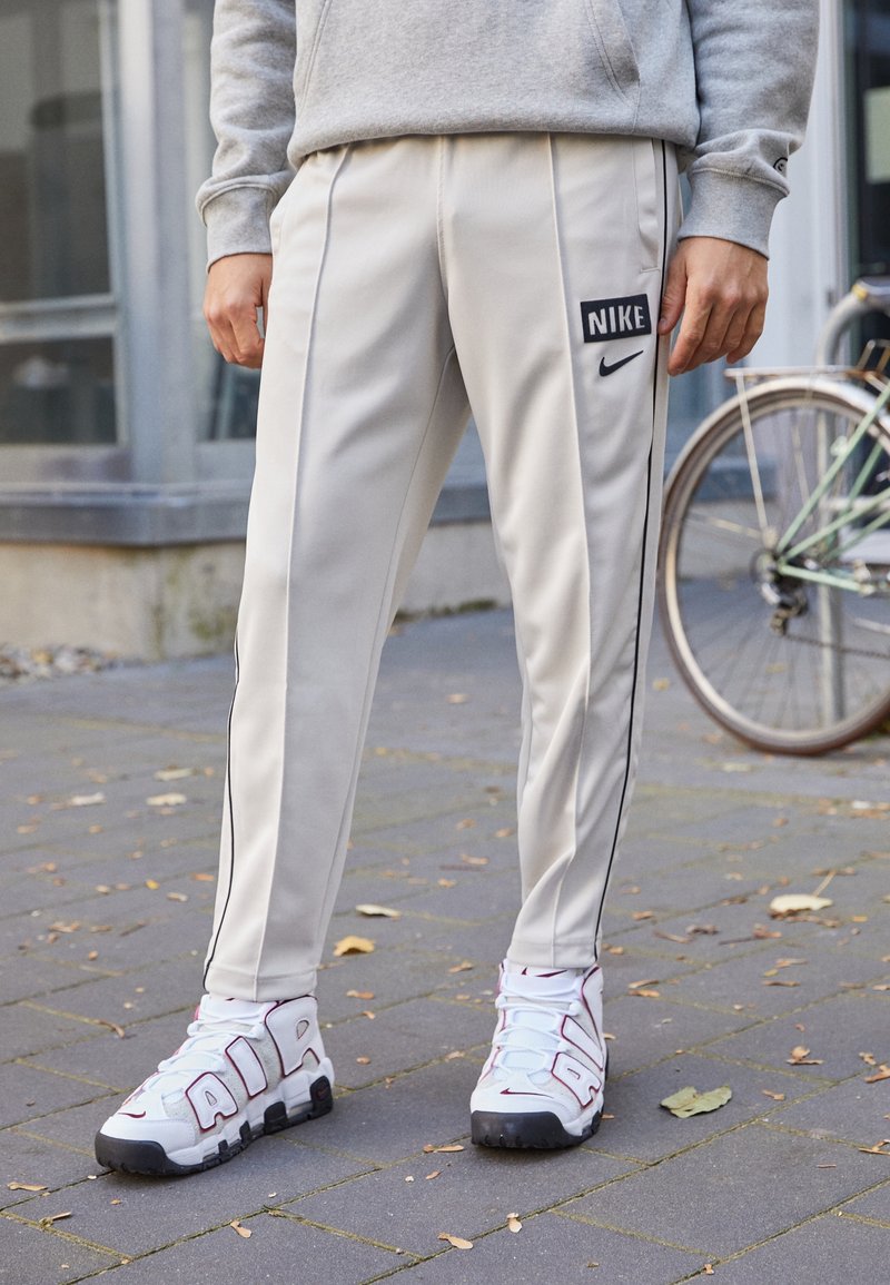 Ljusgrå Nike-träningsbyxor med svarta sidsränder och logotyp. Bärs med vita sneakers med framträdande "AIR"-bokstäver och röda detaljer.