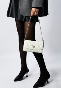 Borsa a tracolla bianca trapuntata con dettagli dorati, tracolla a catena e chiusura a pattina. Il modello indossa un cappotto grigio, una gonna nera plissettata e tacchi neri.