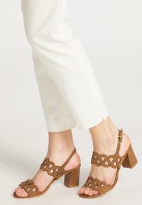 Sandale maro din suede cu design decupat, vârful deschis și toc masiv. Curea ajustabilă la gleznă. Asociate cu pantaloni albi conici.