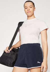 Lyserosa t-skjorte med korte ermer og "Guess" logo, sammen med marineblå joggeshorts med hvit kant og elastisk midje. Svart treningsbag i hånden.