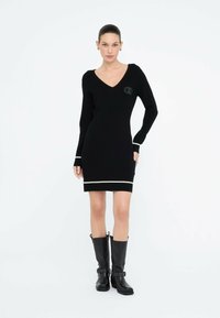 Donna in piedi che indossa un abito lungo a maniche nere con scollatura a V e bordo bianco ai polsini e all'orlo, con stivali neri fino a metà polpaccio con fibbie.