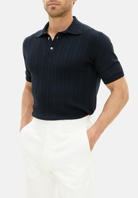 Mörkblå stickad polo med ribbad textur, korta ärmar och platt krage, i kombination med vita skräddarsydda byxor. Tvåknappsdetalj på knäppningen.