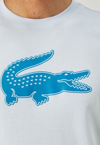 White cotton t-shirt featuring a large blue crocodile graphic with triangular patterns on its back and detailed facial features.