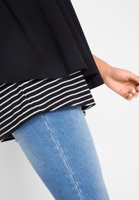 Haut noir ample superposé à une chemise rayée blanche et noire, associé à un jean skinny bleu clair. Tissu lisse et design décontracté.