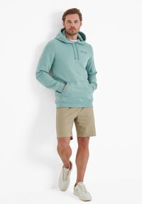 Light teal hoodie with front pouch, adjustable drawstring, and embroidered logo. Paired with beige shorts and white sneakers.