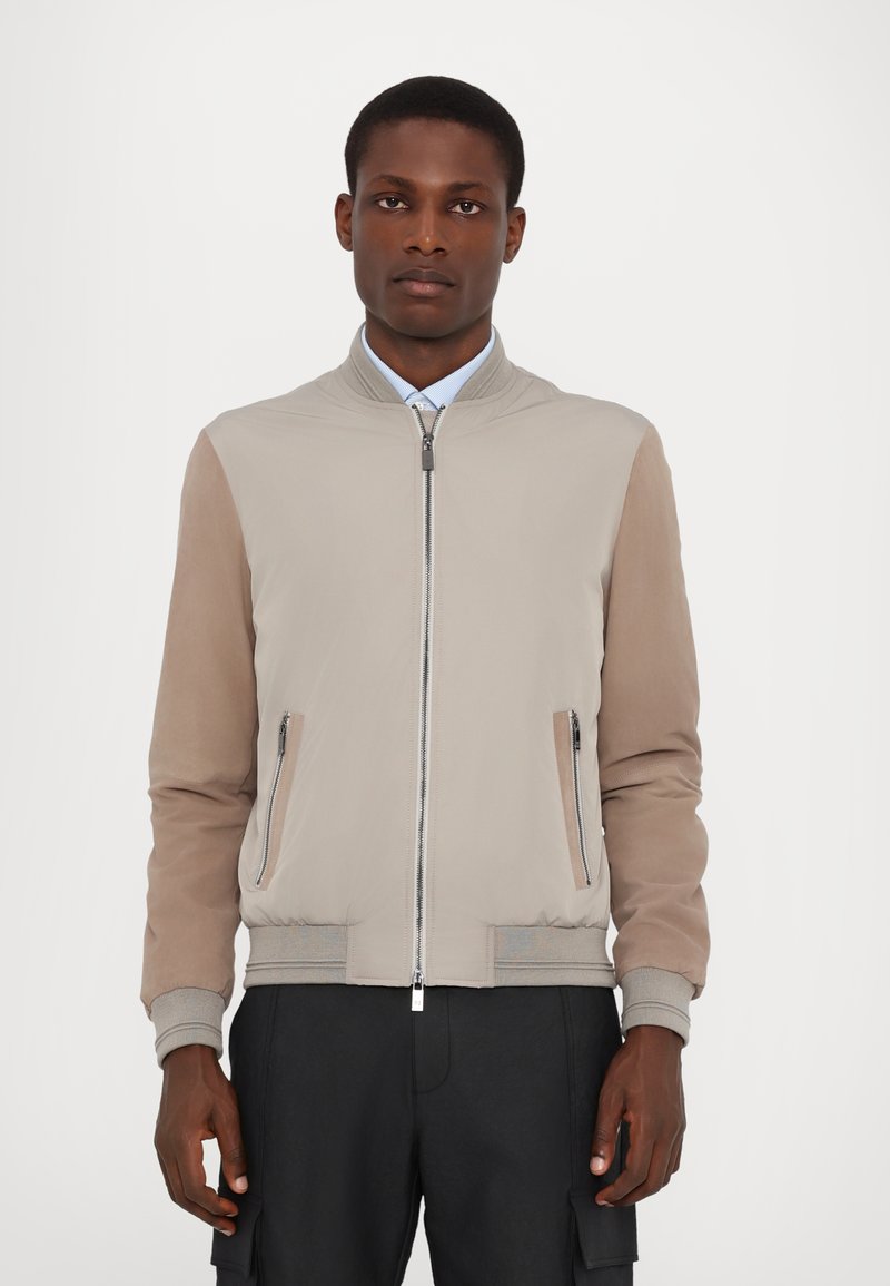 Hellbeige Bomberjacke aus glattem Stoff, mit kontrastierenden braunen Ärmeln, silbernem Reißverschluss und zwei Seitentaschen. Rippbündchen und Saum.