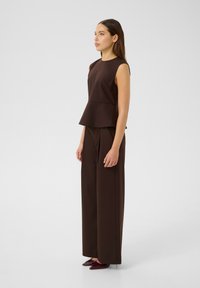 Smeđa majica bez rukava s peplum donjim dijelom, uparena sa širokim smeškim hlačama. Outfit ima glatku teksturu i kroj prilagođen figuri.