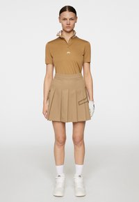 Brun kortärmad polo med mönstrad krage, i kombination med en veckad beige kjol med sidficka och diskret logodetalj. Vita sportskor fulländar looken.