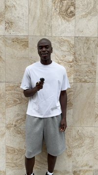 T-shirt branca de manga curta e calções cinzentos, segurando um pequeno objeto preto, de pé contra uma parede de pedra clara texturizada.