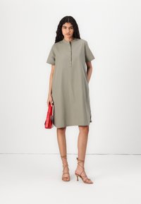 Kratka rukav, olivno zelena obleka z zaobljenim ovratnikom in detajli gumbov spredaj. V kombinaciji z svetlobradimi čeveljčki na peto in rdečo torbico.