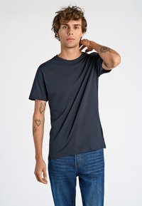 Camiseta azul marino hecha de una tela suave, con un corte relajado, cuello redondo y mangas cortas. Presenta tatuajes en los brazos del modelo.