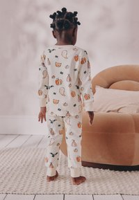 Bomuldspyjamas med en cremefarvet base og farverige mønstre af spøgelser, græskar og flagermus. Ribstrikkede manchetter og tætsiddende design. Blød tekstur.