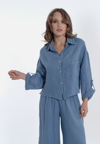 Camicia di lino azzurro chiaro con davanti a bottoni, colletto e taschino. Abbinata a pantaloni coordinati larghi.