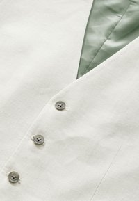 Gilet en lin beige clair avec une doublure intérieure en satin vert foncé. Il dispose de cinq boutons gris, de revers crantés et d'une surface en tissu texturé.
